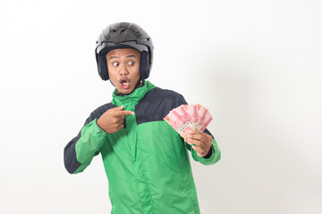 Wall Mural - Portrait of Asian online taxi driver wearing green standing against white background, holding a bunch of rupiah money while pointing to the side