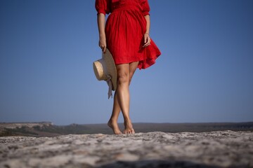 Wall Mural - A woman in a red dress is standing on a rocky beach with a straw hat in her hand