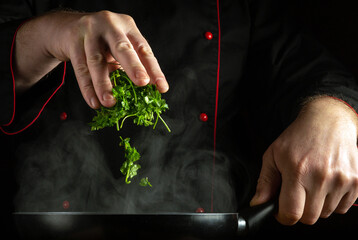 Wall Mural - A professional chef manually adds fragrant fresh parsley to a hot frying pan. The concept of preparing a delicious dinner in a restaurant kitchen.