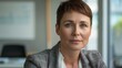 © iuricazac - A professional woman with short hair wearing a blazer sitting in an office with a window in the background.