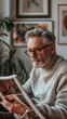 © iuricazac - A man with gray hair and a beard wearing glasses and a white sweater reading a book in a cozy room with framed pictures on the wall.