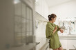 © eggeeggjiew - Asian Woman in green shirt checking smartwatch in modern kitchen, displaying curiosity and anticipation.