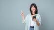 © Jirawatfoto - Excited asian girl shopping on mobile app, holding phone and pointing finger left at empty copy space, showing promo text, standing with smartphone against green background