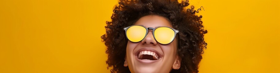 A cheerful young woman with curly hair happily stands against a bright yellow background, wearing trendy reflective sunglasses, exuding joy and a carefree attitude in a vibrant and positive manner