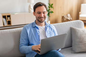 Wall Mural - A man is sitting on a couch while using a laptop. He appears focused on the screen, typing and clicking. The room is well-lit, with a coffee table in front of him.