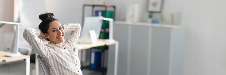 Canvas Print - Happy relaxed lady with hands behind her head enjoying rest from work in office interior, sitting at workplace and smiling at camera. Successful business and great deal, copy space