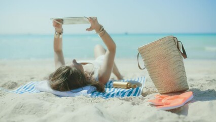 Wall Mural - relaxed elegant 40 years old woman on the seacoast with straw bag and striped towel digital tablet blank screen.