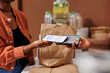 © DC Studio - African American woman uses contactless transaction to purchase organic produce at local market with a mobile phone. Closeup of black woman using nfc payment at checkout counter.