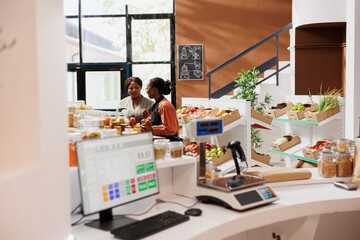 Wall Mural - In eco friendly store, vendor with an apron assisting customer in selecting fresh bulk products with plastic free packaging. Black women standing in bio food shop, talking about sustainable lifestyle.