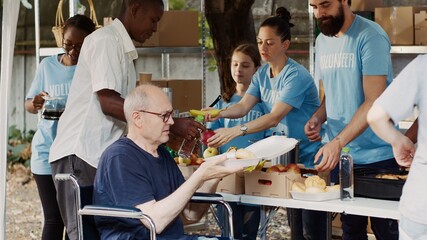 Senior old man in wheelchair receiving free food from group of multiethnic volunteers at hunger relief event. Friendly charity workers providing warm meals to the homeless and disabled people.