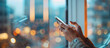 © Asad - Close up of hands holding a phone and searching on the screen, with a blurred office background and blurred window showing light from outside. In the distance is a blurred business center building