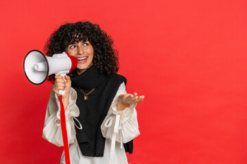 Caucasian young woman with curly hair talking with megaphone, proclaiming news, loudly announcing advertisement discounts sale, using loudspeaker to shout speech. Girl isolated on red background