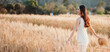 © Satori Studio - A woman is walking through a field of tall golden wheat. She is wearing a white dress and a straw hat. The scene is peaceful and serene, with the woman enjoying the beauty of the natural landscape