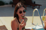 Fototapeta  - Beautiful brunette eating fruit on a stick by a pool