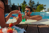 Fototapeta  - Man in a swimming pool toasting a colorful drink to a woman lying on a towel