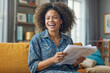 © polack - Beautiful happy Afro woman sits on a sofa in living room and reading a document or letter with good news. Good news, approval of a bank loan, promotion at work