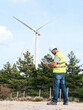 © Koldo_Studio - A technician in a high-visibility vest and hard hat uses a laptop while standing in an open field near wind turbines.