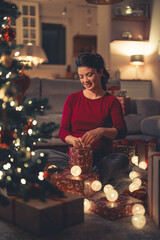 Wall Mural - A beautiful happy young adult woman wrapping Christmas presents while sitting on the ground in a living room