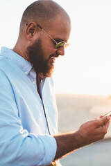 Fototapete - Hipster guy in sunglasses watching online video from networks on smartphone app satisfied with connection, handsome young man reading news browse web page on modern telephone resting on sunny weekend