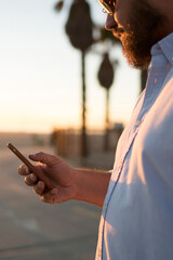 Fototapete - Cropped view of hipster guy holding modern mobile phone in hand reading news from browsed web page, young man sending text message on smartphone using 4G internet connection standing outdoors