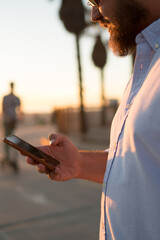 Fototapete - Young bearded guy watching video from social networks using mobile phone 4G connection standing on street, cropped view of male hipster checking notification of modern application holding smartphone
