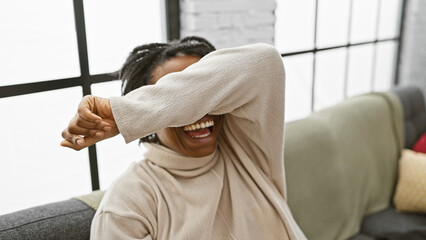 Poster - Laughing young black woman covering eyes with arm indoors, living room background, casual style, joy, playful, expressive, adult