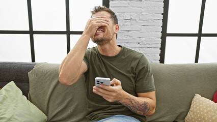 Wall Mural - A smiling man with a beard and a tattoo reacts to his smartphone in a modern living room setting.