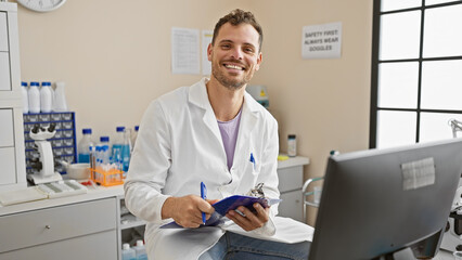 Wall Mural - Handsome hispanic man with beard in a clinic wearing lab coat holding clipboard smiling at camera