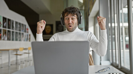 Poster - Young hispanic man in an office reacts joyfully in front of a laptop wearing headphones