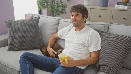 Sticker - Relaxed young hispanic man petting a cat while sitting on a couch indoors with a mug in his hand.