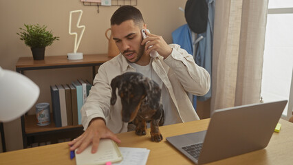 A young hispanic man in casual attire works from home, talking on the phone while his dachshund walks by on the desk near a laptop.