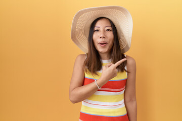 Wall Mural - Middle age chinese woman wearing summer hat over yellow background surprised pointing with finger to the side, open mouth amazed expression.