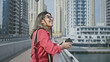 © Krakenimages.com - Smiling woman holding smartphone enjoys sunny dubai marina skyline view