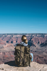 Wall Mural - Back view of young woman with rucksack sitting on rocky cliff enjoying trip in USA exploring wild nature, female traveler fascinated with scenery of landscape with stone formation in National park