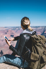 Wall Mural - Rear view of female traveler checking connection on telephone sitting on high mountain hill, hipster girl wanderlust satisfied with data network in wild environment holding smartphone for chatting