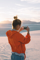 Wall Mural - back view of young woman using smartphone camera for making picture of sunset in white sand dunes at