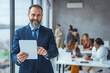 © Dragana Gordic - Portrait of a mature businessman using a digital tablet in a modern office with his colleagues in the background. Cropped shot of a mature businessman working on his tablet in the office