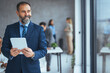 © Dragana Gordic - Happy middle aged business man ceo wearing suit standing in office using digital tablet. Smiling mature businessman professional executive manager looking away thinking working on tech device.