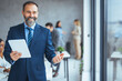 © Dragana Gordic - Smiling mid aged business man wearing suit standing inside office holding digital tablet. Mature businessman professional holding fintech device looking away thinking or new business ideas solutions