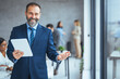 © Dragana Gordic - Smiling mid aged business man wearing suit standing inside office holding digital tablet. Mature businessman professional holding fintech device looking away thinking or new business ideas solutions