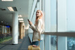 © Natee Meepian - Young woman with suitcase using smartphone at airport terminal. Modern travel concept, casual outfit, waiting for flight