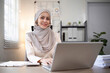 © Natee Meepian - Smiling Muslim woman working on laptop at office desk. Professional workspace, casual attire