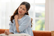 © 220 Selfmade studio - A woman experiencing neck pain while sitting on a couch, showing discomfort. She has her hand on her neck, indicating where the pain is.