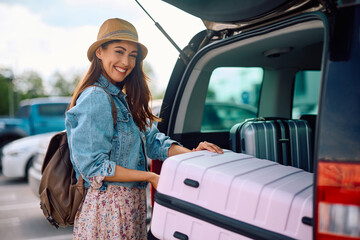 Wall Mural - Happy traveler taking her suitcase out of car trunk on parking lot and looking at camera.