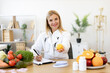 © sofiko14 - Portrait of smiling female nutritionist doctor sitting at table with vegetables and fruits in clinic. Positive mature Caucasian female weighing lemon while writing healthy food recipe on clipboard.