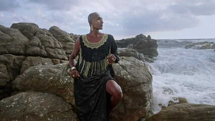 Wall Mural - Non-binary black person in rich dress, golden earrings, nose ring, bracelet, rings sits on stone against stormy ocean waves. Queer lgbtq pretty fashion model wears jewelry in posh gown.