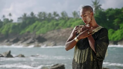 Wall Mural - Trans sexual ethnic fashion model in long posh dress and accessories in elegant posture looks at camera slow motion. Epatage gay black man in luxury gown poses with hands crossed on scenic ocean beach