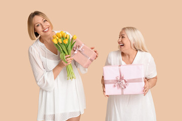 Poster - Adult woman greeting her mother with bouquet of tulips and gift boxes on beige background. International Women's Day celebration