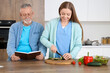 © Pixel-Shot - Senior man reading book with nurse cooking in kitchen