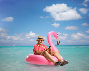 Poster - Mature male tourist sitting on a famingo swimming ring in the sea and using a mobile phone